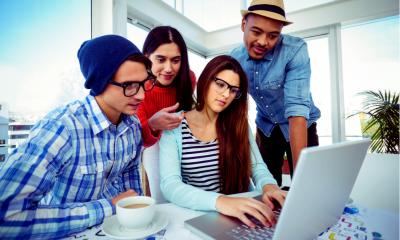 Group of employees dressed in casual work clothes gathered around a laptop