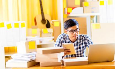 Man in blue and white plaid shirt sat at a desk with a laptop and boxes in front of him