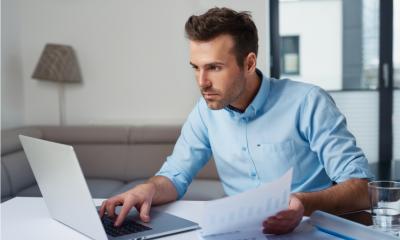 A man sitting a a desk typing on a computer holding a piece of paper