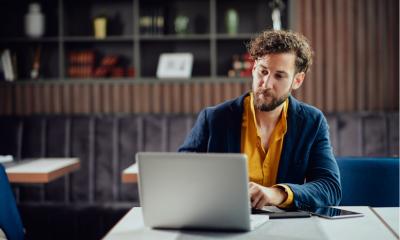 Young serious bearded businessman in smart casual clothes sitting in cafe and typing on laptop.