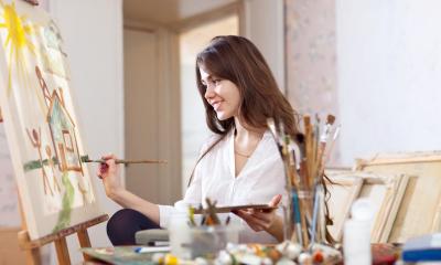 Woman sitting down painting on canvas with paint brushes in foreground