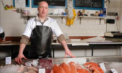 Fishmonger in black apron stood behind table with various fish types on it