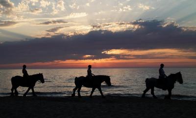 Three people trekking on ponies along a beach with the sunset in background
