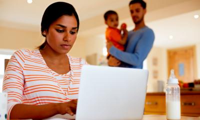 Mum working from home while dad is holding a baby in the background