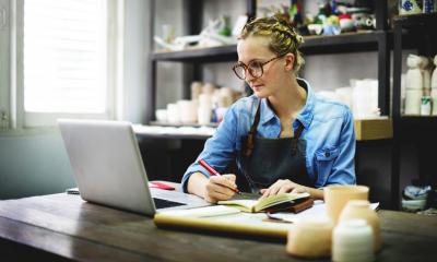 Woman in blue shirt and black apron writing in a notebook and looking at a laptop