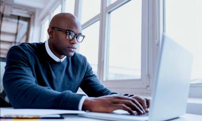 Man in a blue sweater wearing glasses sat at a desk using his laptop for an e-learning tutorial