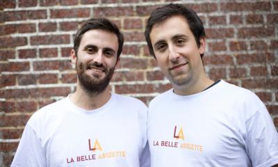 Two men stood in front of a wall with white t-shirts on saying La Belle Assiette