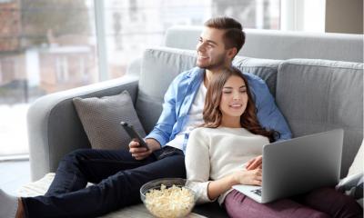 Couple watching TV and whilst woman multitasks using her laptop on a sofa at home