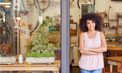 Young mixed race woman smiling while standing in the door of her cafe with her arms folded proud to be the owner of a small business