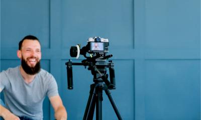 A bearded man shoots video content of himself using camera on tripod.